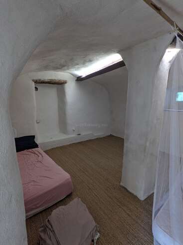 Dieses Bild zeigt ein minimalistisches Zimmer mit weißen, geschwungenen Gipswänden, einem einfachen Bett mit rosa Laken, einem strukturierten Bodenbelag, natürlichem Licht und einem Moskitonetz, das ein weiteres Bett bedeckt.