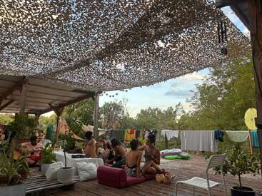 A group of people relax together on a wooden deck under a camouflage canopy, surrounded by plants and laundry, enjoying a casual, outdoor, communal atmosphere.