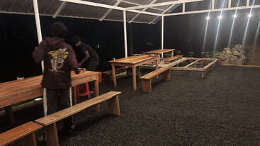 Two people are arranging items on wooden tables and benches under a roofed outdoor area at night, with string lights creating a cozy, inviting atmosphere.