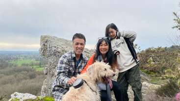 Das Bild zeigt eine dreiköpfige Familie, die mit ihrem Hund auf einem felsigen Hügel vor einem wolkenverhangenen Hintergrund posiert. Die Familie ist leger gekleidet.