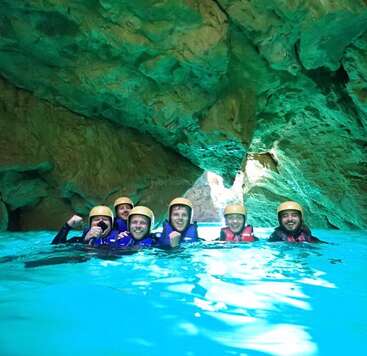 A imagem mostra seis pessoas em uma caverna, usando coletes salva-vidas e capacetes, flutuando em água azul brilhante, com uma grande formação rochosa ao fundo.