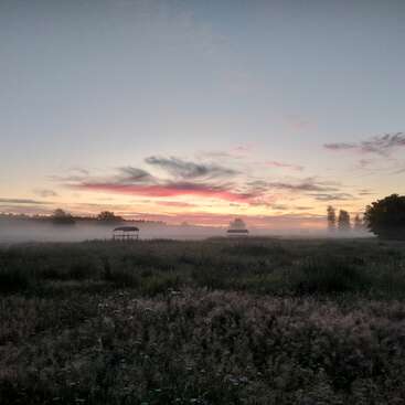Das Bild zeigt eine ruhige Landschaft mit hohem Gras, einer entfernten Scheune und einem farbenfrohen Sonnenuntergang, der ein Gefühl von Frieden und Ruhe hervorruft.