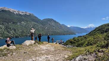 Eine Gruppe von Menschen steht auf einem Hügel mit Blick auf einen wunderschönen blauen See, der von üppig grünen Bergen umgeben ist, unter einem klaren, sonnigen Himmel. Eine atemberaubende Landschaft!