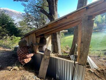 Zwei Leute bauen draußen einen kleinen Schuppen aus Holz und Metall, umgeben von Bäumen und Bergen, und arbeiten gemeinsam in einer malerischen ländlichen Gegend mit grünem Gras in der Nähe.