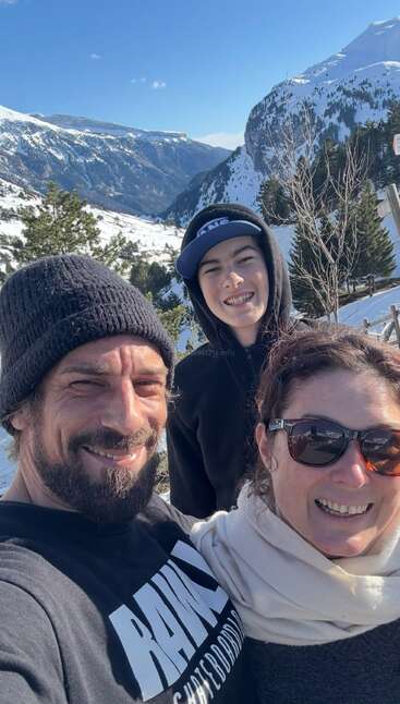 Tres personas sonríen al aire libre con las montañas nevadas y los árboles como telón de fondo. Visten ropa de abrigo y disfrutan del sol y de un impresionante paisaje invernal durante su aventura juntos.