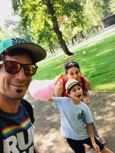 Das Bild zeigt eine dreiköpfige Familie, die für ein Selfie in einem Park posiert. Der Vater trägt eine Sonnenbrille und ein Regenbogenhemd, während der Sohn Zuckerwatte in der Hand hält.