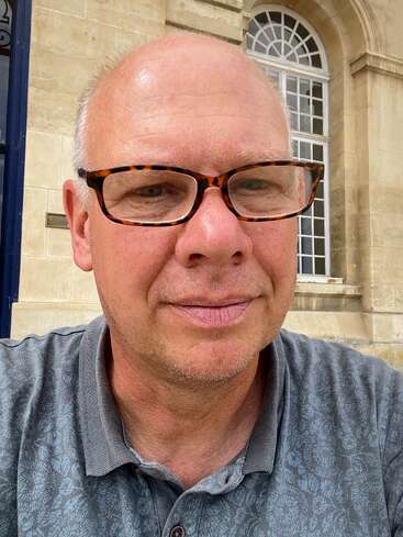 A middle-aged man with glasses and a hint of a smile takes a selfie outside a beige stone building with arched windows, wearing a patterned polo shirt.