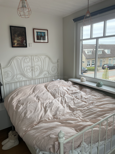 A cozy bedroom with a rumpled pale pink duvet, white metal bed, slippers, two framed pictures, large window, crystal decor, and street view outside.