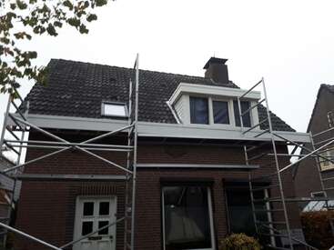This image shows a brick house with a sloped, tiled roof. Scaffolding surrounds the house, indicating renovation or construction work in progress on the roof area.