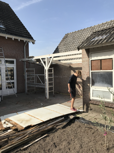 A person is working outside between two brick houses, surrounded by construction materials, wooden boards, scaffolding, and ladders on a clear, sunny day.