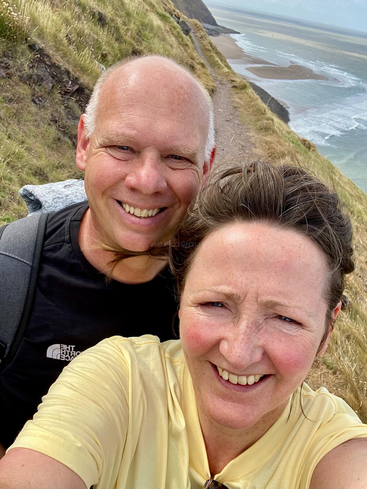 A smiling couple takes a selfie on a scenic coastal hiking trail. Behind them, grassy cliffs and the ocean create a stunning, windswept backdrop under blue skies.