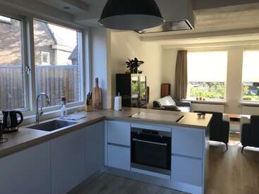 This image shows a modern, bright kitchen connected to a cozy living area. Large windows, wooden countertops, and contemporary furnishings create a welcoming, functional open-plan space.