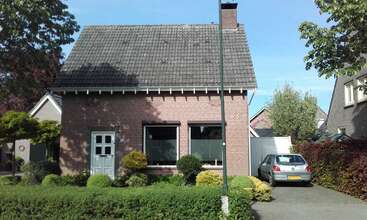 This image shows a brick house with a gray tiled roof and large windows. There’s a small garden, trees, a driveway, and a parked car.