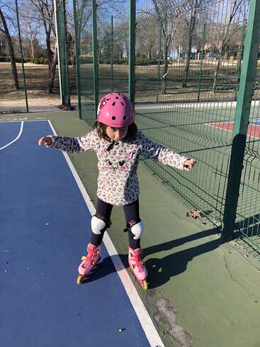 A young girl wearing a pink helmet and rollerblades is skating on a blue and green outdoor court, with a fence and trees in the background.