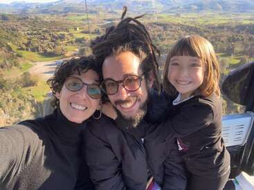 The image depicts a family of three posing for a selfie on a scenic overlook, with rolling hills and mountains visible in the background.