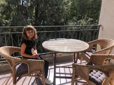 The image depicts a young girl sitting at a small, round table on a balcony, surrounded by wicker chairs and a metal railing, with trees visible in the background.