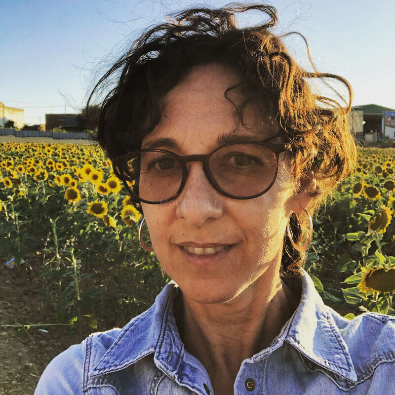 The image depicts a woman with dark hair and glasses standing in front of a field of sunflowers, wearing a denim shirt and smiling at the camera.