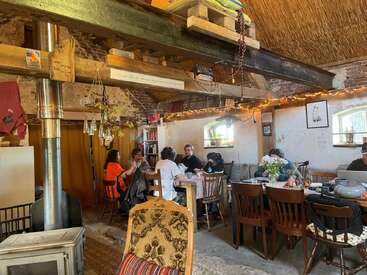 A cozy, rustic room with wooden beams and brick walls. Several people sit around tables eating and talking. Warm lighting, books, flowers, and decorations create a homey atmosphere.