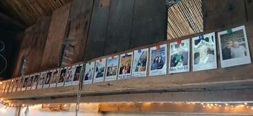 A row of instant photo prints is pinned to a wooden beam, displaying various people and moments. String lights hang below, creating a warm, nostalgic atmosphere.
