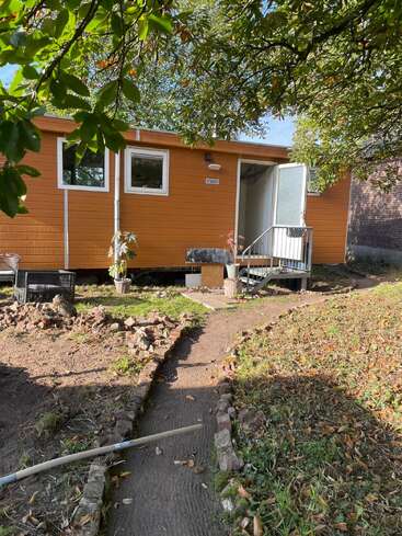 A small orange cabin with white trim, surrounded by trees and greenery. A dirt path, lined with bricks, leads to the open front door.
