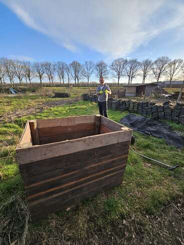 A man stands in a grassy field holding gloves, near stacked boards and a wooden compost bin, with leafless trees and a small shed in the background.