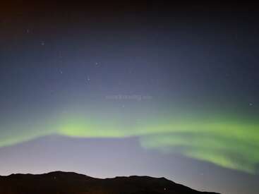 A dark, starlit sky features beautiful green Northern Lights over silhouetted hills. The natural aurora glows softly, creating a mesmerizing, peaceful scene beneath the night sky.