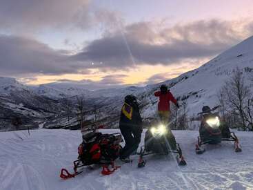 Two people stand with snowmobiles on a snowy mountain at sunset. The sky glows purple and orange. Snowy peaks and a winding valley surround them.