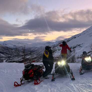 Deux personnes se tiennent debout avec des motoneiges sur une montagne enneigée au coucher du soleil. Le ciel brille de couleurs violettes et orangées. Des sommets enneigés et une vallée sinueuse les entourent.
