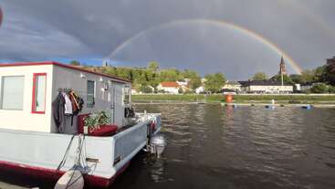 Una casa flotante está atracada en un río con ropa secándose fuera, bajo un vibrante arco iris doble, con árboles verdes y casas visibles en la orilla opuesta.