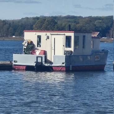 Uma pequena casa flutuante com telhado vermelho flutua em um lago calmo, cercada por árvores e uma costa distante, sob um céu parcialmente nublado à luz do dia.