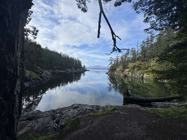 A calm, clear inlet surrounded by forested rocky shores, reflected in the still water, under a bright blue sky with scattered clouds. Peaceful and serene nature.