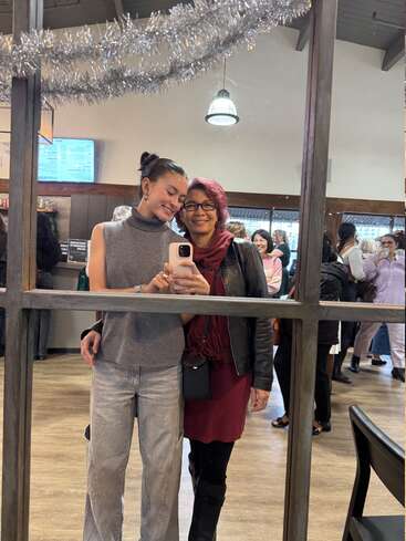 A young woman and an older woman smile together for a mirror selfie, surrounded by festive decorations in a busy, warmly lit café filled with people.