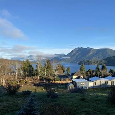 A peaceful lakeside village with scattered houses, trees, and open fields. Majestic mountains and serene water are beautifully illuminated under a clear blue sky.