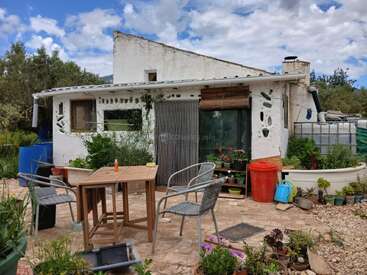 Une maison blanche rustique au décor éclectique, entourée de plantes, de meubles d'extérieur et de récipients colorés, sous un ciel partiellement nuageux dans un cadre naturel.