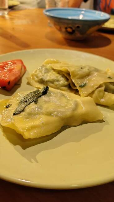 Três pedaços de ravióli cobertos com sálvia, servidos em um prato amarelo com uma fatia de tomate, em uma mesa de madeira. Um ambiente aconchegante para o jantar.