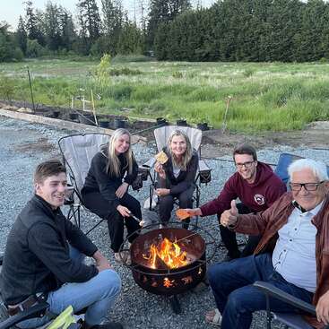 Das Bild zeigt eine Gruppe von fünf Personen, die um eine Feuerstelle versammelt sind, Marshmallows rösten und die Gesellschaft der anderen in einer ruhigen Umgebung genießen.