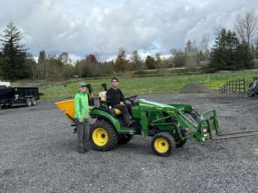Two people are outdoors on a gravel lot with a green John Deere tractor. One stands beside it, the other is seated, surrounded by rural scenery.