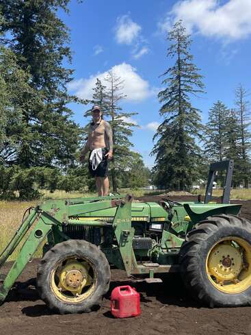 A shirtless man stands on an old green John Deere tractor in a rural field, surrounded by tall trees, under a bright blue sky with clouds.