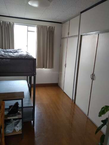 A small, tidy bedroom with bunk beds, wooden floor, cream-colored curtains, built-in closets, a desk, and a single potted plant in the corner.