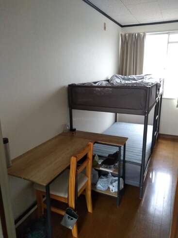 This image shows a small, tidy room with a metal bunk bed, a wooden desk, a chair, shelves with items, and a window with curtains.