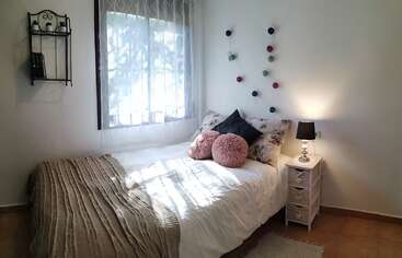 This cozy bedroom features a neatly made single bed with decorative pillows, a small nightstand with a lamp, wall art, window, and a shelf.