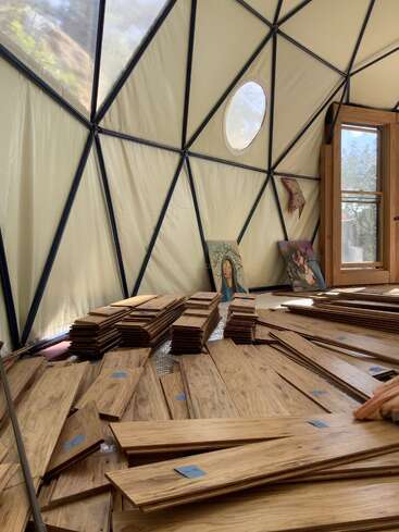 Im Inneren einer geodätischen Kuppel mit dreieckigen Fenstern bedecken ordentlich gestapelte Holzbretter den Boden. Zwei Gemälde und einige Wandkunstwerke lehnen an den Stoffwänden.