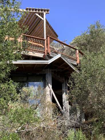 Eine rustikale Holzhütte liegt inmitten von dichtem Grün. Sie verfügt über eine geräumige Terrasse mit Holzgeländer und einer Pergola mit Lattenrost, unter einem klaren blauen Himmel.