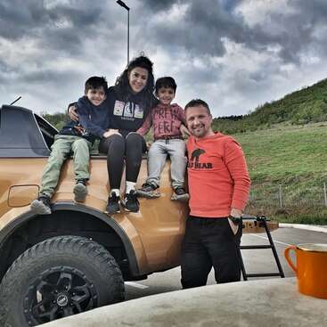 L'image représente une famille de quatre personnes posant à l'arrière d'un camion orange, avec un ciel nuageux et une colline herbeuse en arrière-plan.