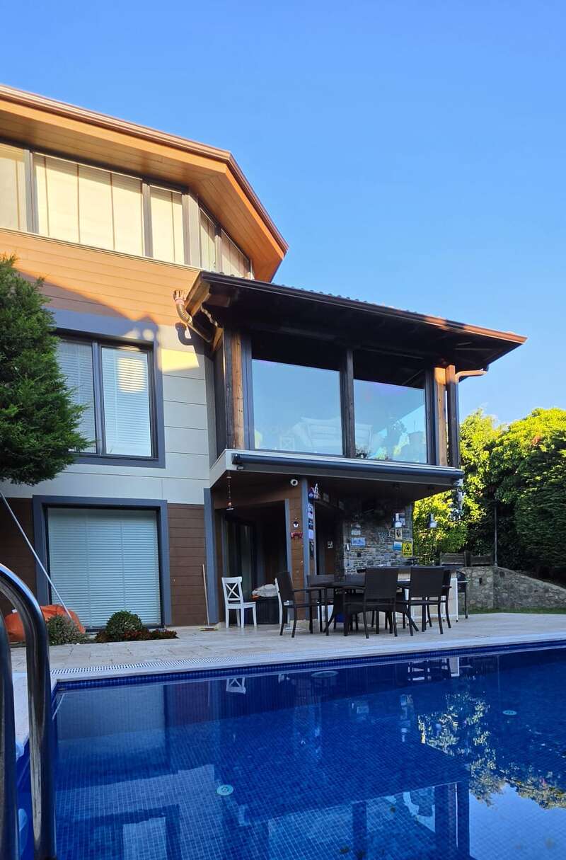 Une maison moderne dotée de grandes baies vitrées donne sur une piscine bleue. On y trouve un coin salon en plein air et de la verdure tout autour, sous un ciel bleu clair et lumineux. Un cadre paisible.