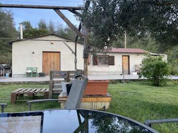 A rustic house with wooden doors sits in a lush garden. Outdoor furniture, a table, plants, and trees create a peaceful rural atmosphere on a sunny day.