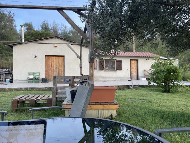 A rustic house with wooden doors sits in a lush garden. Outdoor furniture, a table, plants, and trees create a peaceful rural atmosphere on a sunny day.