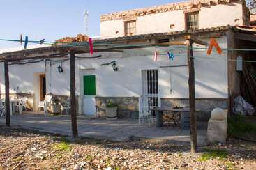 A rustic country house with white walls, green door, outdoor patio furniture, clotheslines with colorful pegs, potted plants, and a partially tiled roof under blue sky.