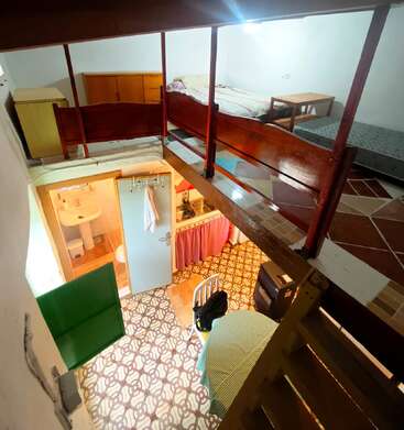 This cozy loft apartment features a small upstairs bedroom with a bed and dresser, overlooking a compact living area, kitchenette, dining table, and a bathroom below.