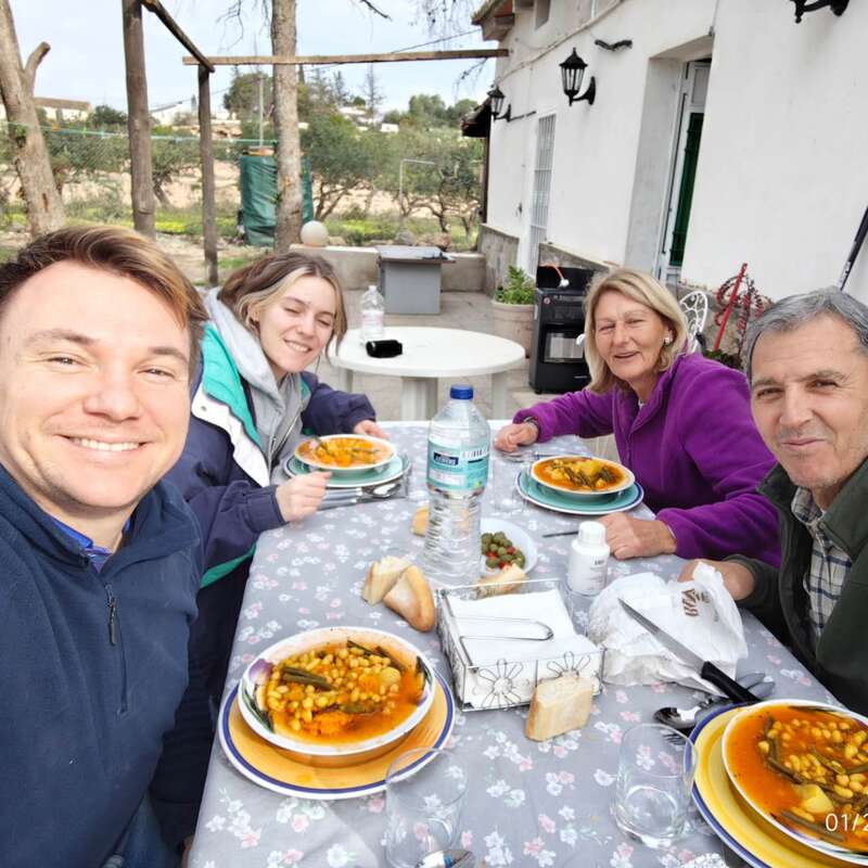 Four people are enjoying a meal together outdoors. They are smiling, sitting around a table with dishes of soup, bread, water, and a pleasant atmosphere.